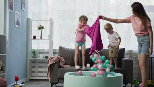 Loving mother with daughter and son pours colorful balls from bag into dry pool. Toddler boy plays with Rubik cube. Happy family has fun together at home