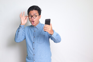 Portrait of shocked Asian man in blue shirt looking at mobile phone. Wow face expression. Isolated image on white background