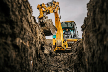 A worker in bulldozer is moving soil and making hole on construction site. - obrazy, fototapety, plakaty