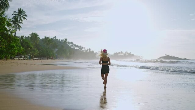 Young blonde woman jogs by sea at sunrise. Caucasian female runs on beach by ocean at dusk. Fit sporty girl exercises outdoor with tropical background. Healthy body lifestyle. Back view tracking shot