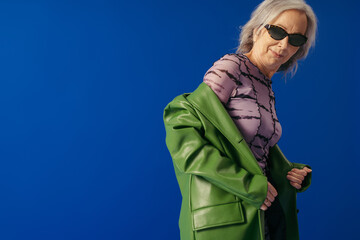 trendy senior woman posing in sunglasses and green leather jacket over lilac dress isolated on blue