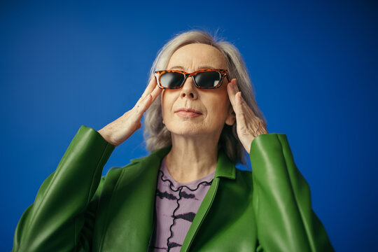 elderly woman in green leather jacket adjusting trendy sunglasses isolated on blue