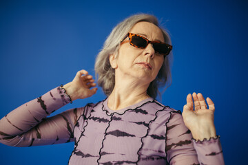 senior woman in stylish sunglasses posing while looking away isolated on blue