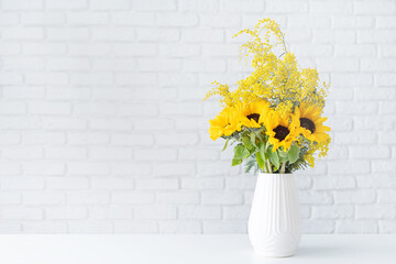 Fototapeta na wymiar Blossom of Mimosa and sunflowers in white ceramic vase on white brick wall background with space for text