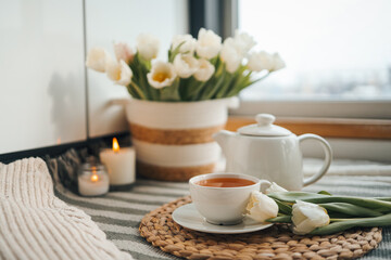 Cup of tea, teapot, basket with tulips, candles in home interior, aesthetic photo