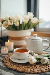 Cup of tea, teapot, basket with tulips, candles in home interior, aesthetic photo