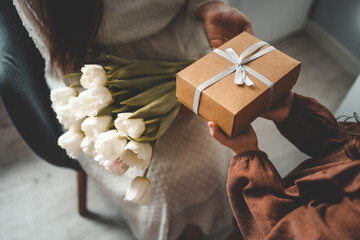 The child's hands hold a beautiful gift box with a ribbon and white tulips. Top view, close-up. Happy mother's day - obrazy, fototapety, plakaty