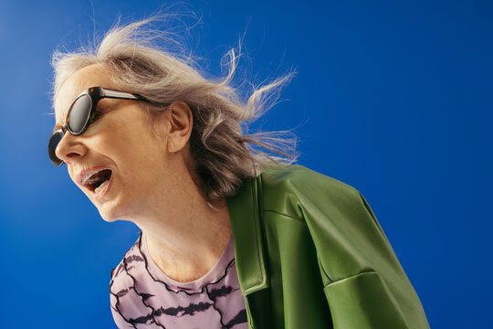 senior woman in trendy sunglasses and green leather jacket standing with opened mouth against wind isolated on blue