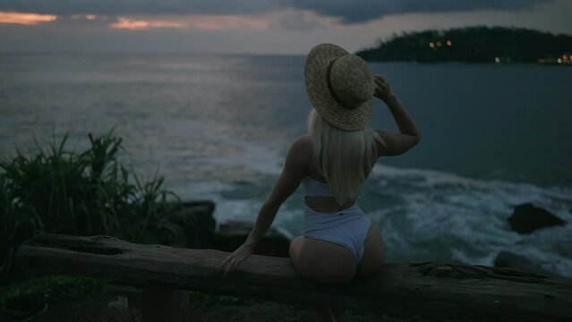 Back view of sexy woman in swimwear and straw hat sitting on bench on edge of picturesque hill with sea view and lights on background after sunset. Slim blonde curvy female sits seductively in dark