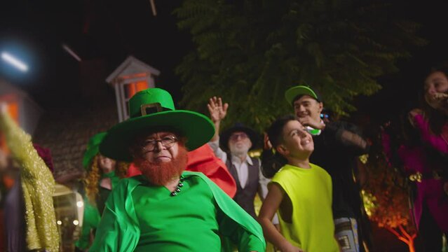 Group of people dancing , having fun outside at the night . Old fat man wearing leprechaun costume , dances funny dance. Celebrating Halloween or Saint Patrick's day . Crowd dances on house background