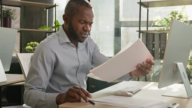 Nervous tired African American middle-aged mature man ethnic businessman has problem with business documents mistake read wrong paper loan bills feel headache sit at office desk sunbeams background