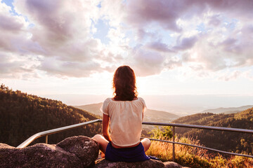 Frau meditierend bei Sonnenuntergang auf einem Berg mit Weitblick und strahlendem Gegenlicht - obrazy, fototapety, plakaty