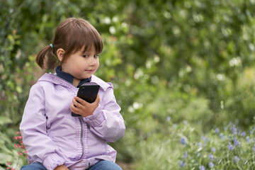 A little girl is chatting on a smartphone in a blooming garden. She has a surprised or incredulous...