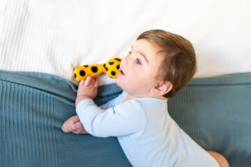 baby on bed with light blue onesie and big blue eyes, blue-gray blanket. little baby playing with yellow stuffed giraffe, top view with copy space