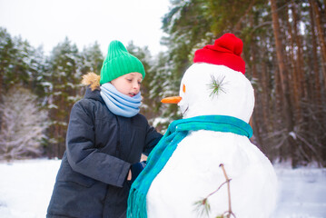 Fototapeta na wymiar the boy made a snowman, a happy child plays in a snowy winter forest, straightens a scarf and a hat for a snowman