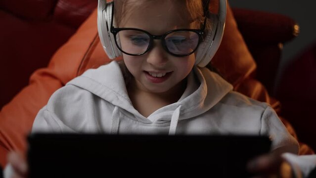 Excited teen girl plays game at home on digital tablet technology device sitting on sofa. Emotional child in headphones and glasses holds pad computer surfing internet.Children tech addiction concept.