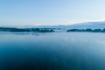 Foggy morning on a beautiful lake surrounded by forests and fields. A small village located near a lake in fog and morning sun.