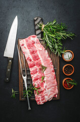 Raw pork ribs with rosemary and spices, meat fork and chef knife on rustic wooden cutting board prepared for cooking on black kitchen table background, top view