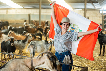 Happy young Latin female traveler in plaid shirt waving Peruvian flag during visit to livestock goat farm