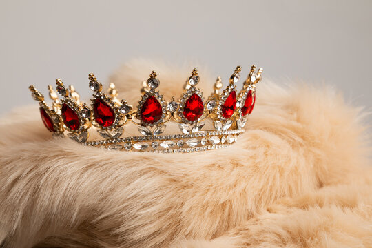 Close up product photo of a golden crown with red jewels. The crown is on a pink fur rug.