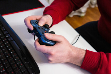 Side view of male hands hold the joystick and presses the buttons in front of the computer screen. Player plays a video game using controller at home. Boy gamer playing racing videogame. Closeup.