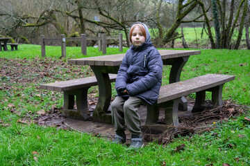 A young boy is sitting on a bench with a table to relax. Green grass around. The boy is wearing a warm jacket.