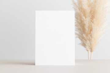 White invitation card mockup with a pampas decoration on the beige table. 5x7 ratio, similar to A6, A5.