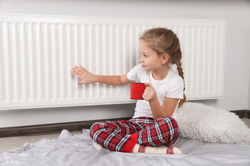 Little girl with cup of hot drink near heating radiator indoors