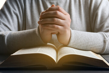 Woman holding hands clasped while praying over Bible at wooden table, closeup