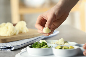 Baby food. Woman putting piece of cauliflower into section plate with different vegetables at grey textured table, closeup