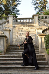 Portrait of beautiful female model with blonde plait, wearing black leather catsuit and hooded cloak, fantasy assassin warrior. Standing pose holding archery bow and arrow weapon, castle background.