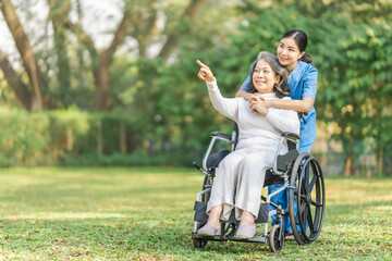 Young asian care helper with asia elderly woman on wheelchair relax together park outdoors to help and encourage and rest your mind with green nature. Hand pointing forward