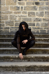 Portrait of beautiful female model with blonde plait, wearing black leather catsuit and flowing hooded cloak, fantasy assassin warrior.  Posing in castle background with stone staircase.