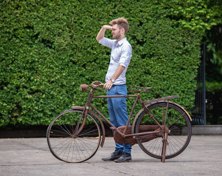 Man in white shirt blue jeans stand and pose with Old decay bicycle on green vine climbing garden wall outdoor. Rust Classic bike old bicycle on green garden wall retro style.