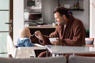 Father wearing bathrope spoon feeding hir infant baby boy child sitting in high chair at the dining table in kitchen at home in the morning.