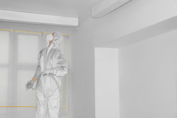 Decorator in protective overalls painting ceiling with spray gun indoors