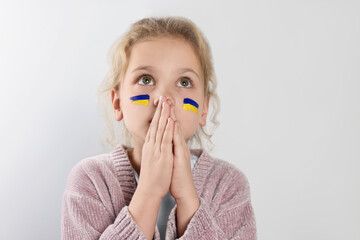 Little girl with drawings of Ukrainian flag on face and clasped hands against white background