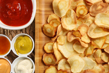 Tasty homemade parsnip chips with different sauces on wicker surface, flat lay