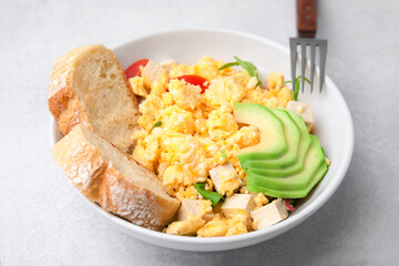 Bowl with delicious scrambled eggs, tofu, avocado and slices of baguette on white table, closeup