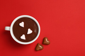 Cup of hot chocolate with heart shaped marshmallows and tasty candies on red background, flat lay. Space for text