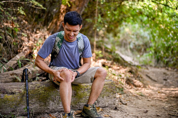 A stressed Asian male trekker sits on a wooden log and holds his knee, suffering from knee pain