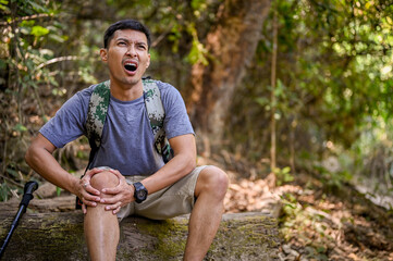 A stressed Asian male trekker sits on a wooden log and holds his knee, severe leg pain, injured