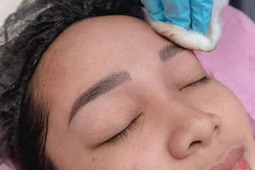 An esthetician wipes off excess pigment and blood from the eyebrows of a customer with a ball of...