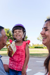 Fototapeta premium Little girl eating banana with parents sitting outdoors on the grass in a park.