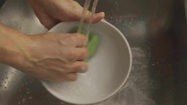 Man hands are wash white soup plate in a metallic sink in close-up