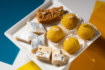 Various Indian desserts (round 'laddu', square beige 'mawa burfi', square brown 'mohanthal', and diamond 'kaju') served on a square white ceramic plate.