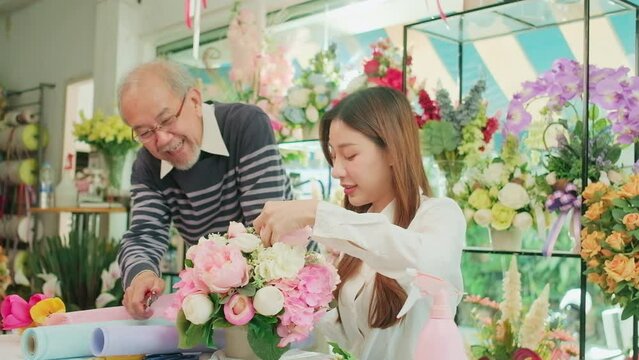 Asian young beautiful female florist worker and senior man shop owner arranging blossoms bouquet together in valentine, happy work with smile and cheerful in colorful flower store, SME entrepreneur. 