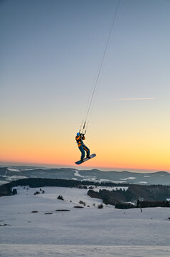 Snowkiting, Snowboarder fliegt mit Zugdrachen durch den Sonnenuntergang am Himmel der Wasserkuppe in der Rh&ouml;n, Hessen, Deutschland