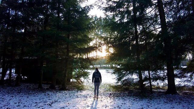 Silhouette of a young woman with a backpack exploring the forest at sunset. Slow motion shot of the entrance to a clearing covered in snow, full of sunlight on a cold winter's day