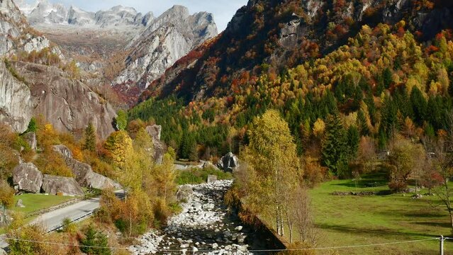 Aerial shot of an autumn landscape with yellow, green and red trees, a rocky river and mountains on the horizon, in Val di Mello in Lombardy, Italy.